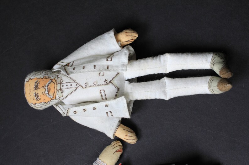 May include: A white fabric doll with a detailed face and a white suit. The doll is lying on a dark surface.