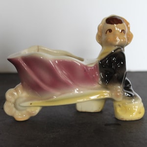 May include: Vintage ceramic planter in the shape of a person pushing a wheelbarrow. The figure has a tan face and hair, a black jacket, and a pink and yellow wheelbarrow. A unique decorative piece.