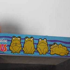 Puede incluir: Una caja con las palabras "Decorative FROGS" en letras rosas y naranjas. La caja presenta cuatro ilustraciones de ranas amarillas en diferentes poses sobre un fondo azul con l&iacute;neas onduladas moradas.