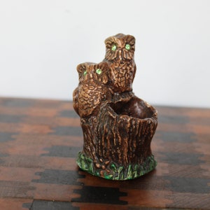 May include: A ceramic owl figurine with green eyes, perched on a tree stump with a small planter. The owl is brown and has a detailed texture.