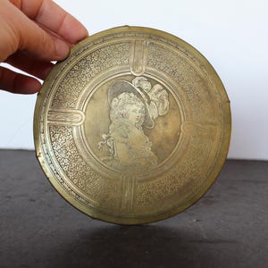 May include: A round, antique brass container with an engraved portrait of a woman in a hat. The container has a detailed floral pattern around the portrait and on the outer rim. The brass has a warm, aged patina.