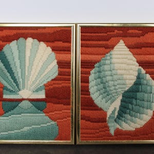 May include: Two rectangular wall art pieces featuring needlepoint shell designs. Each frame contains a textured, stitched seashell in shades of teal, light blue, and cream, set against a vibrant, horizontally striped background of deep orange and red.