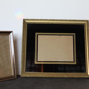 May include: Two gold-toned picture frames. One is rectangular and upright, with a small easel back. The other is a larger, square frame with a black mat and gold trim. Both frames are empty.