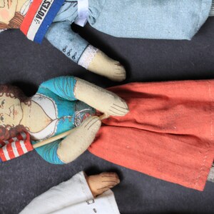 May include: Three vintage fabric dolls with painted faces. One doll is holding a small American flag. Another doll is wearing a blue dress with a white collar and a red skirt. The third doll is wearing a blue and white dress with a red, white, and blue sash that says "I Votes".