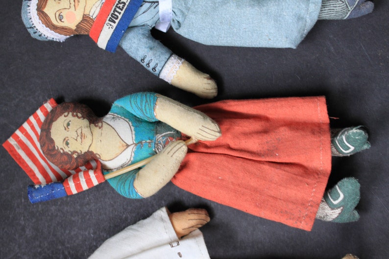 May include: Three vintage fabric dolls with painted faces. One doll is holding a small American flag. Another doll is wearing a blue dress with a white collar and a red skirt. The third doll is wearing a blue and white dress with a red, white, and blue sash that says "I Votes".