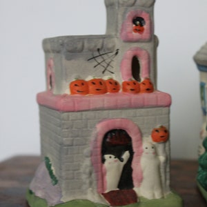 May include: A gray ceramic Halloween house with a pink roof and a pink door. The house has a cobweb design on the side and a row of orange pumpkins on the roof. There are two white ghosts standing in the doorway.