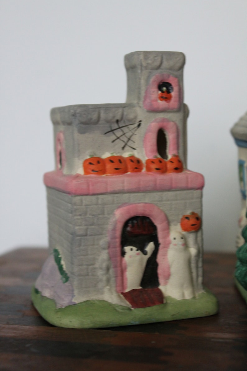 May include: A gray ceramic Halloween house with a pink roof and a pink door. The house has a cobweb design on the side and a row of orange pumpkins on the roof. There are two white ghosts standing in the doorway.