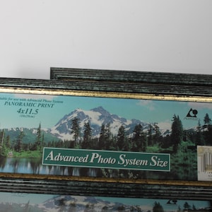 May include: Rectangular picture frames with a dark teal and gold trim. The frames hold a panoramic print of a mountain landscape. The frames are labeled "Advanced Photo System Size" and "4x11.5".