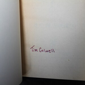 May include: Open book pages with the handwritten name "Tim Colwell" in red ink on the right page. The left page is blank. The pages are off-white, and the book is open on a dark surface.