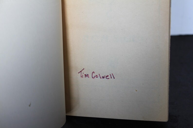 May include: Open book pages with the handwritten name "Tim Colwell" in red ink on the right page. The left page is blank. The pages are off-white, and the book is open on a dark surface.