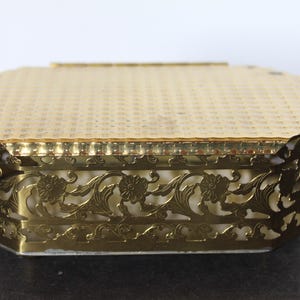 May include: A vintage gold-toned jewelry box with an octagonal shape. The lid has a textured pattern, and the sides feature an ornate floral filigree design. The box is likely made of metal and has a cream-colored interior.