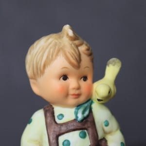 May include: A porcelain figurine of a boy wearing a green and yellow shirt with brown overalls and a green and white scarf. A yellow bird is perched on his shoulder.