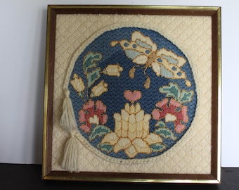 May include: Framed vintage needlepoint art featuring a blue circular design with a butterfly, flowers, and a heart. The artwork is set against a cream-colored background with a diamond pattern, framed in a gold-toned metal frame.