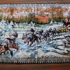 May include: A rectangular tapestry depicting a winter scene with a team of wolves pulling a sled through the snow. A person in a red coat and white hat is on the sled. The border is black with a floral pattern.