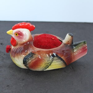 May include: A ceramic pincushion shaped like a hen with a bright red comb, yellow beak, and brown, black, and yellow feathers. The center of the hen's back features a circular, plush, deep red velvet cushion for holding sewing needles and pins.