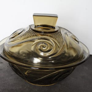 May include: A vintage amber-colored glass serving bowl with a lid. The bowl and lid feature a decorative swirl and textured pattern. The lid has a rectangular handle. The bowl is on a dark surface.