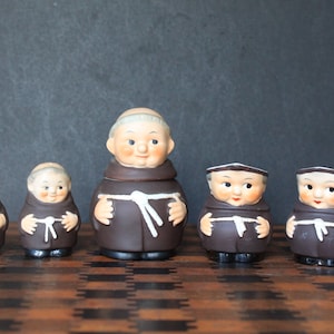 May include: Set of five ceramic figurines of monks, each wearing a brown robe with a white rope belt. The figurines are all smiling and have a whimsical look.