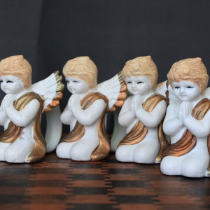 May include: Four white ceramic angel figurines with golden accents, wings, and light brown hair are arranged in a row. Each angel is kneeling with hands clasped in prayer. The background is a dark gray.
