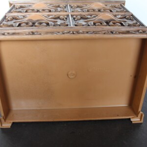 K&ouml;nnte beinhalten: Eine braune dekorative Kunststoffbox mit detailliertem, geschnitztem Deckel. Die Box hat eine rechteckige Form mit einem einfachen, schmucklosen Innenraum. Die Oberseite weist ein verziertes, florales Design auf. Die Box wird von unten betrachtet.