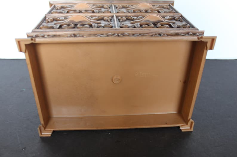 K&ouml;nnte beinhalten: Eine braune dekorative Kunststoffbox mit detailliertem, geschnitztem Deckel. Die Box hat eine rechteckige Form mit einem einfachen, schmucklosen Innenraum. Die Oberseite weist ein verziertes, florales Design auf. Die Box wird von unten betrachtet.