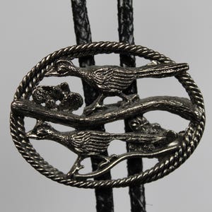May include: Silver-toned bolo tie featuring a detailed oval pendant with two roadrunner birds perched on a branch. The pendant is encircled by a rope-like border and hangs from a black braided cord.
