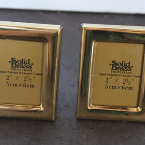 May include: Two rectangular, gold-colored brass photo frames. Each frame has a gold-colored interior and the text "Solid Brass PHOTO FRAME" and measurements of 2" x 2.5" and 5cm x 6cm.