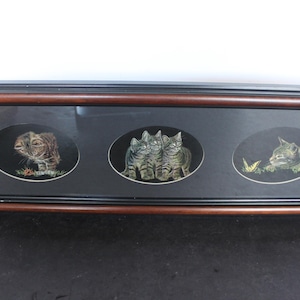 May include: A framed artwork featuring three oval portraits of cats. The cats are depicted in a detailed, realistic style with varying poses and settings. The frame is a dark brown color, and the background of each portrait is black.