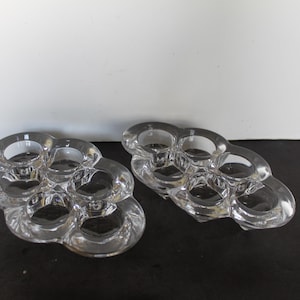 May include: Two clear glass candle holders, each with six circular openings for candles. The holders have a rounded, organic shape and sit on a dark surface. The glass is transparent, allowing light to pass through.