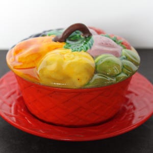 May include: A red ceramic serving dish with a lid featuring a colorful arrangement of sculpted fruits and vegetables. The dish sits on a matching red plate. The lid has a brown handle.