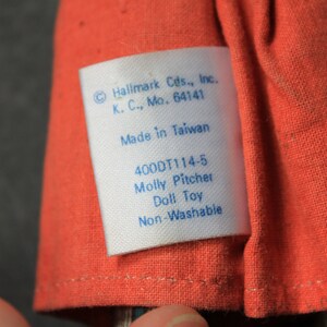 May include: A white tag with blue text attached to a red fabric doll. The tag reads "Hallmark Cds., Inc. K. C., Mo. 64141 Made in Taiwan 400DT114-5 Molly Pitcher Doll Toy Non-Washable".
