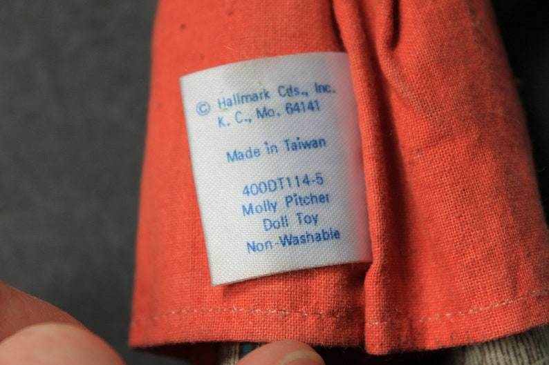 May include: A white tag with blue text attached to a red fabric doll. The tag reads "Hallmark Cds., Inc. K. C., Mo. 64141 Made in Taiwan 400DT114-5 Molly Pitcher Doll Toy Non-Washable".