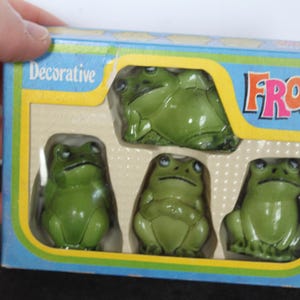 Puede incluir: Un conjunto de figuras decorativas de ranas verdes en una caja azul, amarilla y blanca. La caja dice "Decorative FROGS". Las ranas son brillantes y tienen detalles.