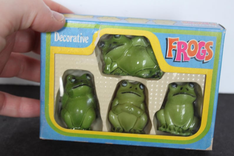 Puede incluir: Un conjunto de figuras decorativas de ranas verdes en una caja azul, amarilla y blanca. La caja dice "Decorative FROGS". Las ranas son brillantes y tienen detalles.
