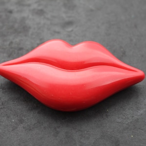 May include: A bright red, glossy, three-dimensional brooch shaped like a pair of full, puckered lips. The object features a smooth, reflective surface with defined curves and a central horizontal line representing the mouth opening.
