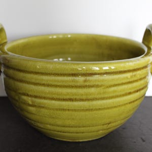 May include: A handcrafted, olive-green ceramic bowl with two loop handles. The bowl features horizontal, textured lines around the exterior. The interior is smooth and glossy. A decorative piece for the kitchen or dining room.