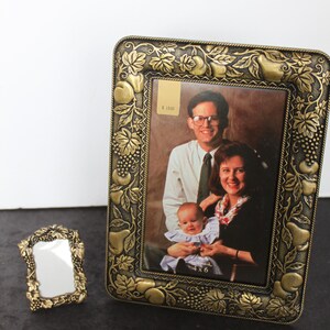 May include: Two decorative picture frames. The larger frame holds a family portrait and has a gold-colored, ornate design with leaves and fruit. The smaller frame is also gold-colored and has a mirror. The larger frame has a price tag.
