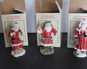 May include: Three boxed figurines of Santa Claus from The International Santa Claus Collection. Each Santa is dressed in a red suit with white trim and a white beard. The Santa on the left is holding a pipe and a toy. The Santa in the middle is holding a goblet. The Santa on the right is holding a staff and a sprig of holly.