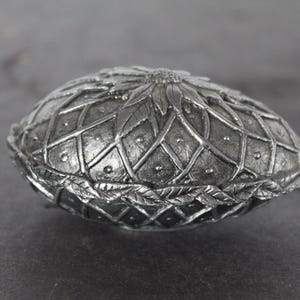 May include: A silver-colored, oval-shaped trinket box with a detailed, textured surface. The lid features a floral design and a lattice pattern. The box is likely made of metal and has a decorative, antique appearance.