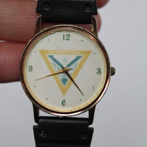 May include: A vintage wristwatch with a gold-toned case and a black band. The watch face is white with black numerals and gold hands. The face features a yellow and blue triangle with the text "INTERNATIONAL MEL CLUB US".
