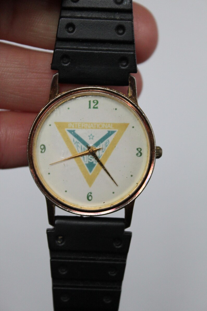 May include: A vintage wristwatch with a gold-toned case and a black band. The watch face is white with black numerals and gold hands. The face features a yellow and blue triangle with the text "INTERNATIONAL MEL CLUB US".