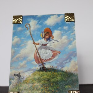May include: A framed print featuring a pastoral scene with a young girl in a bonnet and dress, holding a staff, and a sheep. The artwork is set against a backdrop of a blue sky with clouds and is framed with gold-colored corner accents.