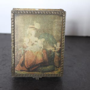 May include: A small, rectangular trinket box with a hinged lid. The box features a framed image of a historical scene with figures in period clothing. The frame is a metallic color with a decorative edge, and the box appears to be made of metal.