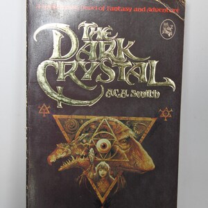 May include: A vintage paperback book titled "The Dark Crystal" by A.C.H. Smith. The cover features gold lettering and a detailed illustration of a creature with a triangle and eye design. The book is dark brown with the text "With 8 Pages of Color Stills from the Magical Movie!"
