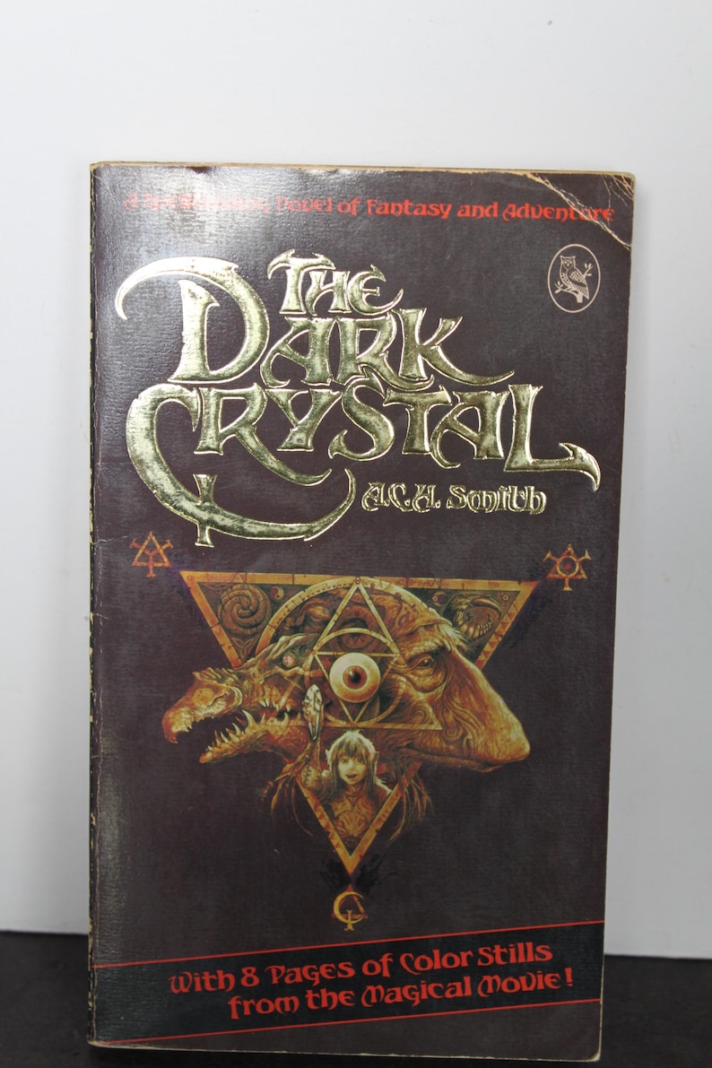 May include: A vintage paperback book titled "The Dark Crystal" by A.C.H. Smith. The cover features gold lettering and a detailed illustration of a creature with a triangle and eye design. The book is dark brown with the text "With 8 Pages of Color Stills from the Magical Movie!"