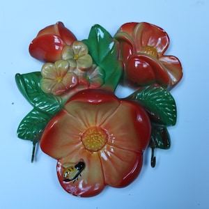 May include: A ceramic floral wall hanging featuring red and orange flowers, green leaves, and a small bee. The flowers have a glossy finish and are arranged in a cluster. Two hooks are attached for hanging.