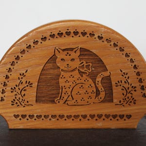 May include: Wooden cat-shaped napkin holder. The holder is a light brown color and features a carved design of a cat with a bow, surrounded by hearts and floral accents. The cat is sitting inside an arched frame.