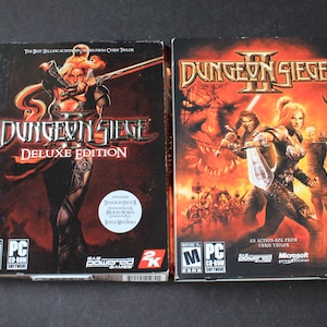 May include: Two PC game boxes for Dungeon Siege II. The left box is the Deluxe Edition and features a female character in armor holding a sword. The right box features a group of characters in a battle scene with a dragon in the background. Both boxes have an M rating.