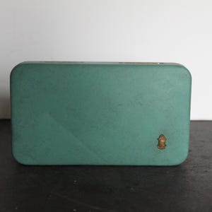 May include: A rectangular, teal-colored box with rounded corners. A small, gold-colored emblem is affixed to the lower right corner. The box is set against a neutral background.