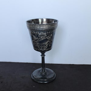May include: A silver-toned metal goblet featuring an intricate engraved band around the rim and a central decorative motif of a banner and foliage. The vessel stands on a slender stem with a circular base, showcasing a polished metallic finish.