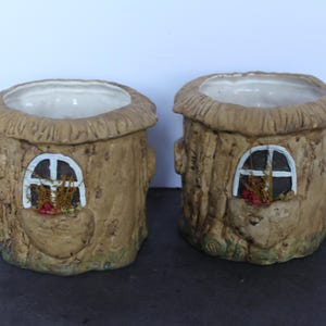 May include: Two ceramic mugs shaped like tree stumps with textured brown bark surfaces. Each mug features a white window frame with small red accents. One mug has a green owl attached to the side, while the other has a yellow raccoon attached to the side.